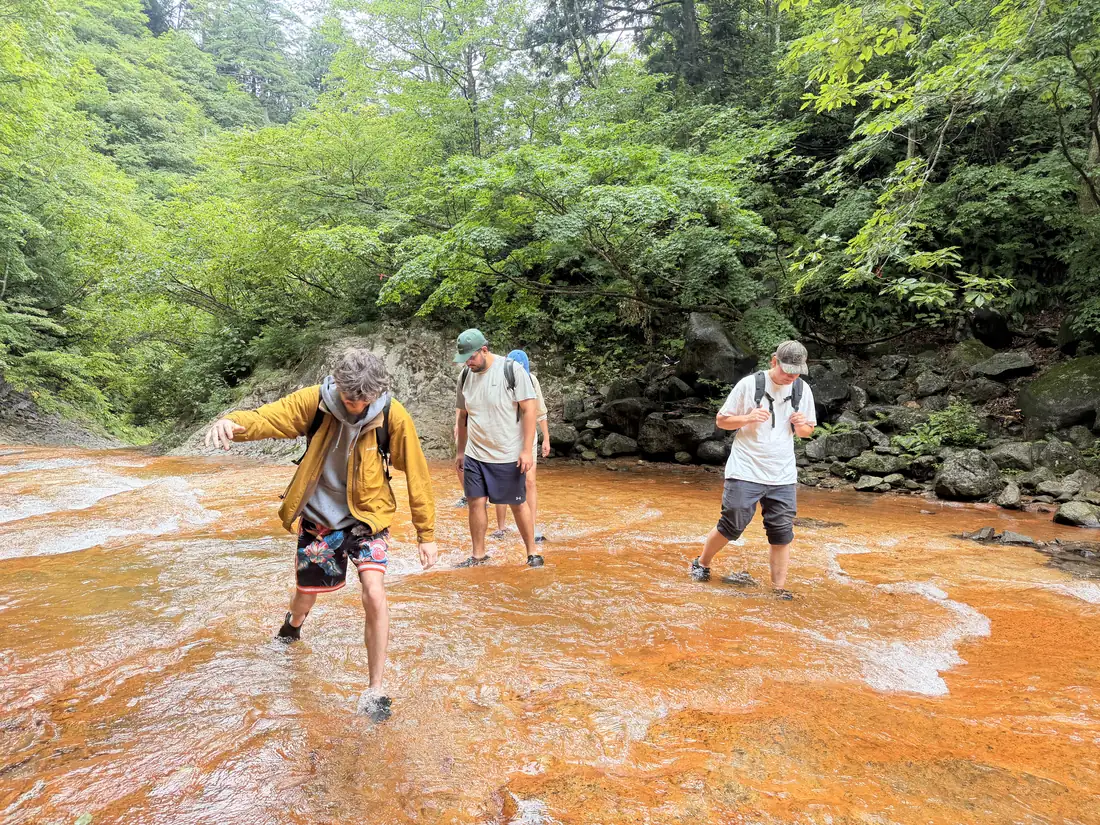 Travelers wade across a vivid orange riverbed on the trail to Namekawa Great Falls