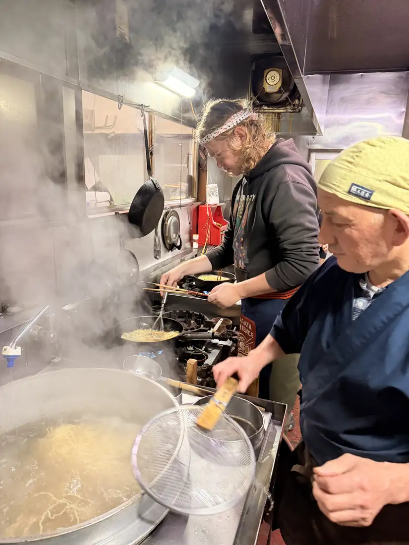 A traveler lifts noodles from a steaming pot beside a village ramen master