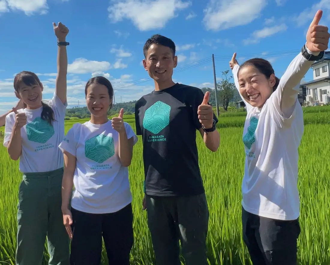 Four YAMAGATA EXPERIENCE guides in logo T-shirts, cheering in front of a green rice field